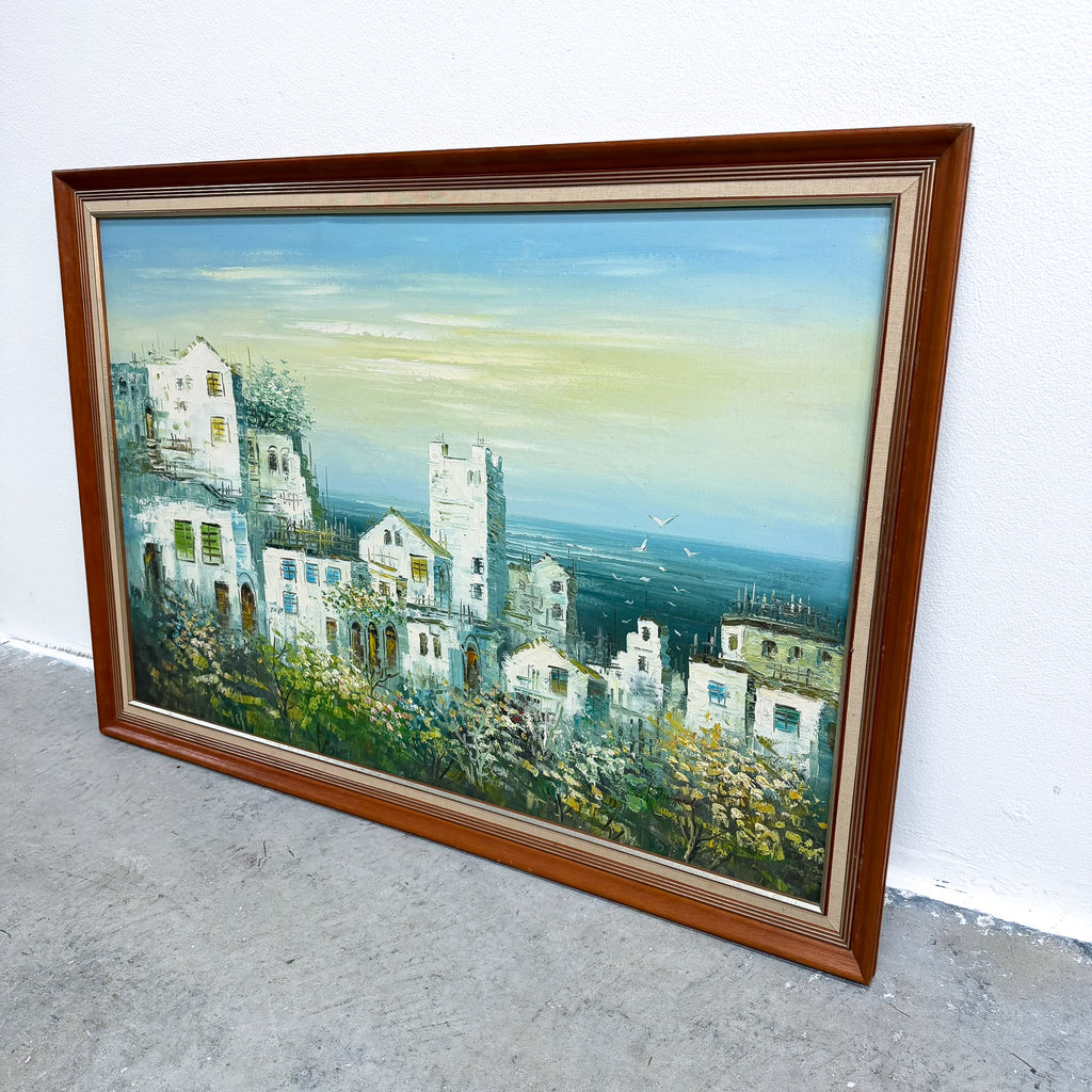 A framed landscape painting of a coastal town with white buildings, greenery, and a pale blue sky, shown leaning against a white wall on a gray concrete floor.