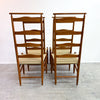 A pair of mid-century modern wooden dining chairs with woven cane seats, shown from the back against a plain white wall and concrete floor.