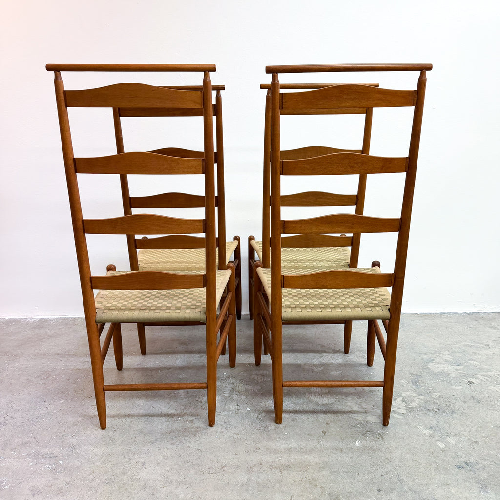 A pair of mid-century modern wooden dining chairs with woven cane seats, shown from the back against a plain white wall and concrete floor.