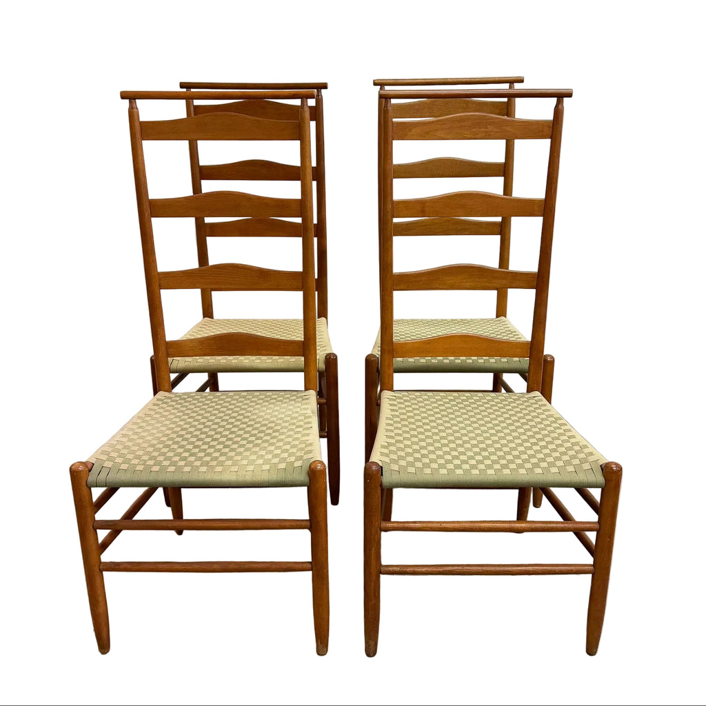 Four wooden dining chairs with woven beige and white checkered seats, arranged in two pairs against a plain white wall and concrete floor.