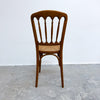 A wooden dining chair with a curved backrest, vertical spindles, and a woven cane seat, photographed against a plain white wall and gray concrete floor.