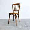 A vintage wooden dining chair with a spindle-back design and a woven seat, positioned against a plain white wall and gray concrete floor.