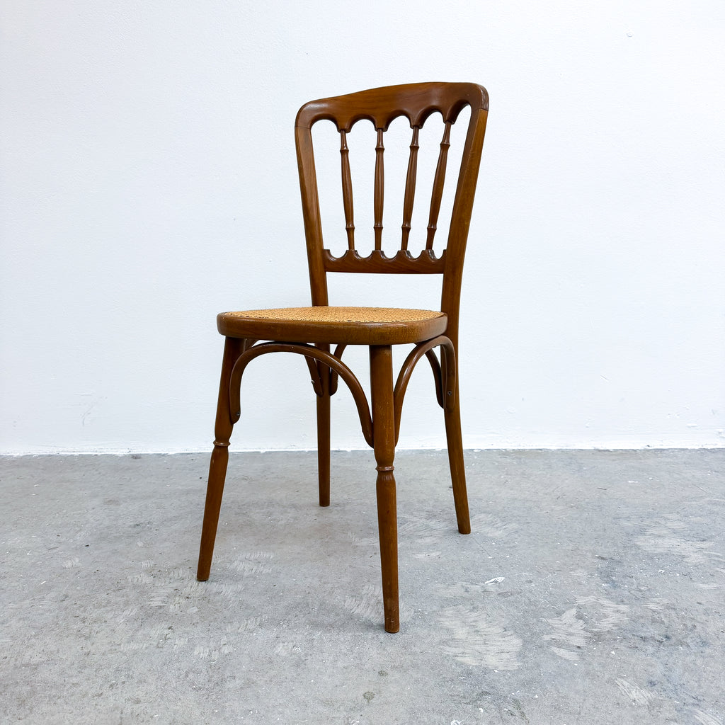 A vintage wooden dining chair with a spindle-back design and a woven seat, positioned against a plain white wall and gray concrete floor.