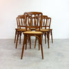 A set of six wooden dining chairs with spindle-back designs and woven cane seats, arranged in a row against a plain white wall and concrete floor.