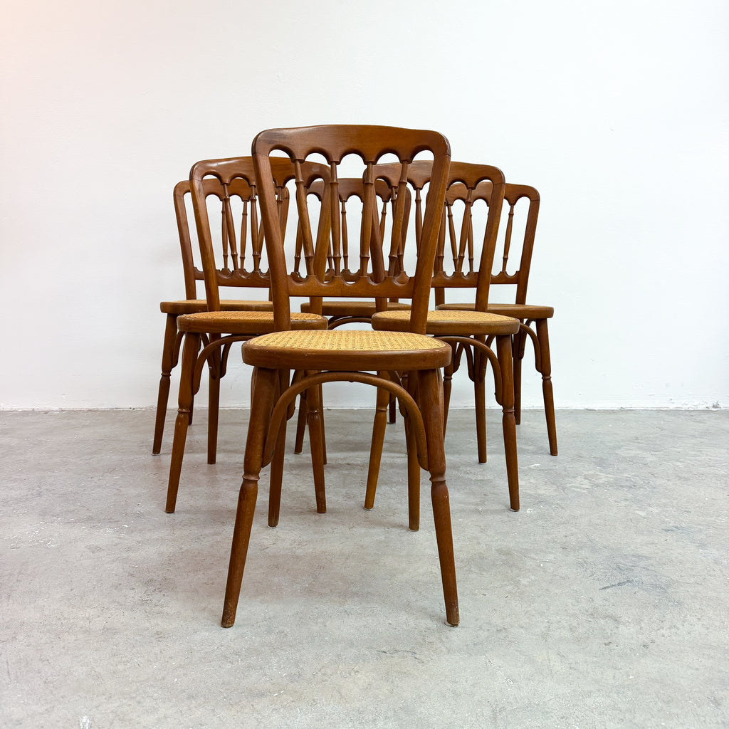 A set of six wooden dining chairs with spindle-back designs and woven cane seats, arranged in a row against a plain white wall and concrete floor.