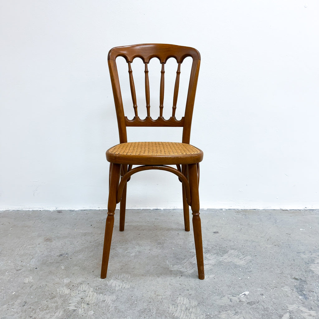 A vintage wooden dining chair with a spindle-back design and a woven cane seat, positioned against a plain white wall and gray concrete floor.