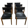 Six mid-century modern wooden dining chairs with black leather seats, arranged in two rows of three on a concrete floor against a white wall.