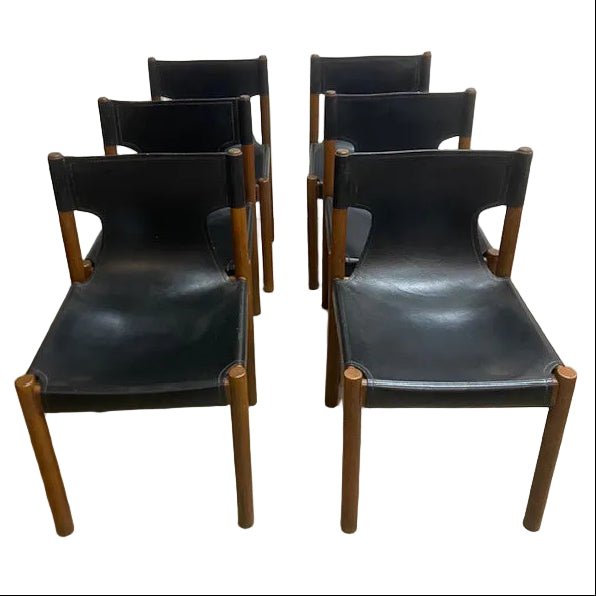 Six mid-century modern wooden dining chairs with black leather seats, arranged in two rows of three on a concrete floor against a white wall.