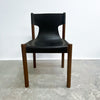 A mid-century modern dining chair with a black leather seat and backrest, supported by a warm brown wooden frame, set against a plain white wall and concrete floor.