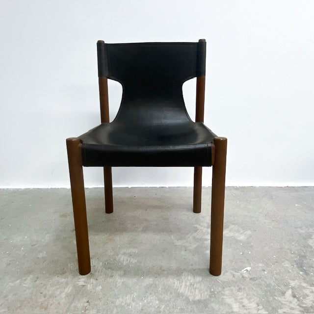 A mid-century modern dining chair with a black leather seat and backrest, supported by a warm brown wooden frame, set against a plain white wall and concrete floor.