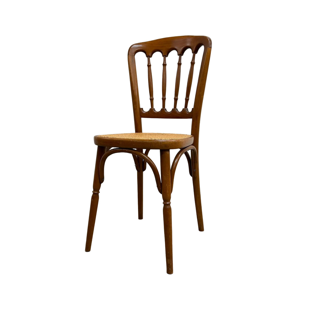 A vintage wooden dining chair with a spindle-back design and a woven seat, positioned against a plain white wall and gray concrete floor.