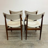 Four mid-century modern dining chairs with dark wood frames and light beige upholstered seats and curved backrests, arranged in two pairs on a concrete floor against a plain white wall.