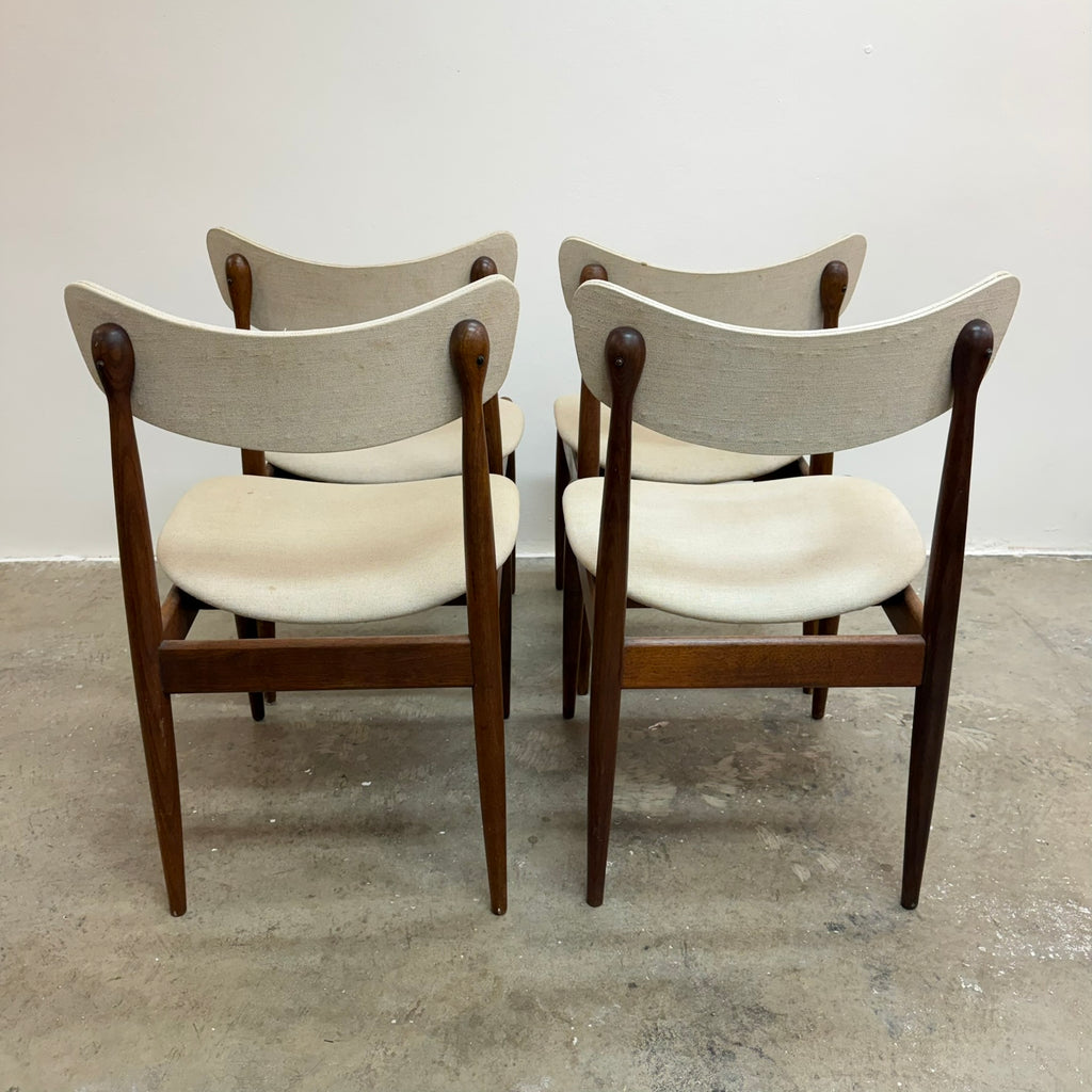 Four mid-century modern dining chairs with dark wood frames and light beige upholstered seats and curved backrests, arranged in two pairs on a concrete floor against a plain white wall.