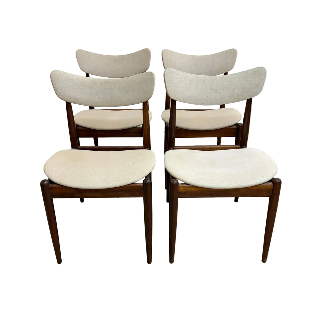 Four mid-century modern dining chairs with curved cream fabric seats and backs on dark wood frames, arranged in two pairs against a plain white wall and concrete floor.