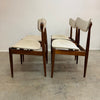 Four mid-century modern wooden dining chairs with cream upholstered seats and curved backrests, arranged on a concrete floor against a plain off-white wall.