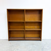 A mid-century modern wooden bookcase with a warm honey-brown finish, featuring six open shelves arranged in a two-column layout, set against a plain white wall and concrete floor.