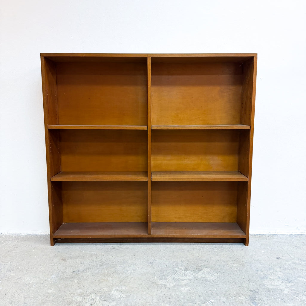 A mid-century modern wooden bookcase with a warm honey-brown finish, featuring six open shelves arranged in a two-column layout, set against a plain white wall and concrete floor.