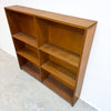 A mid-century modern wooden bookcase with a warm medium-brown finish, featuring six open shelves, photographed against a white wall and concrete floor.