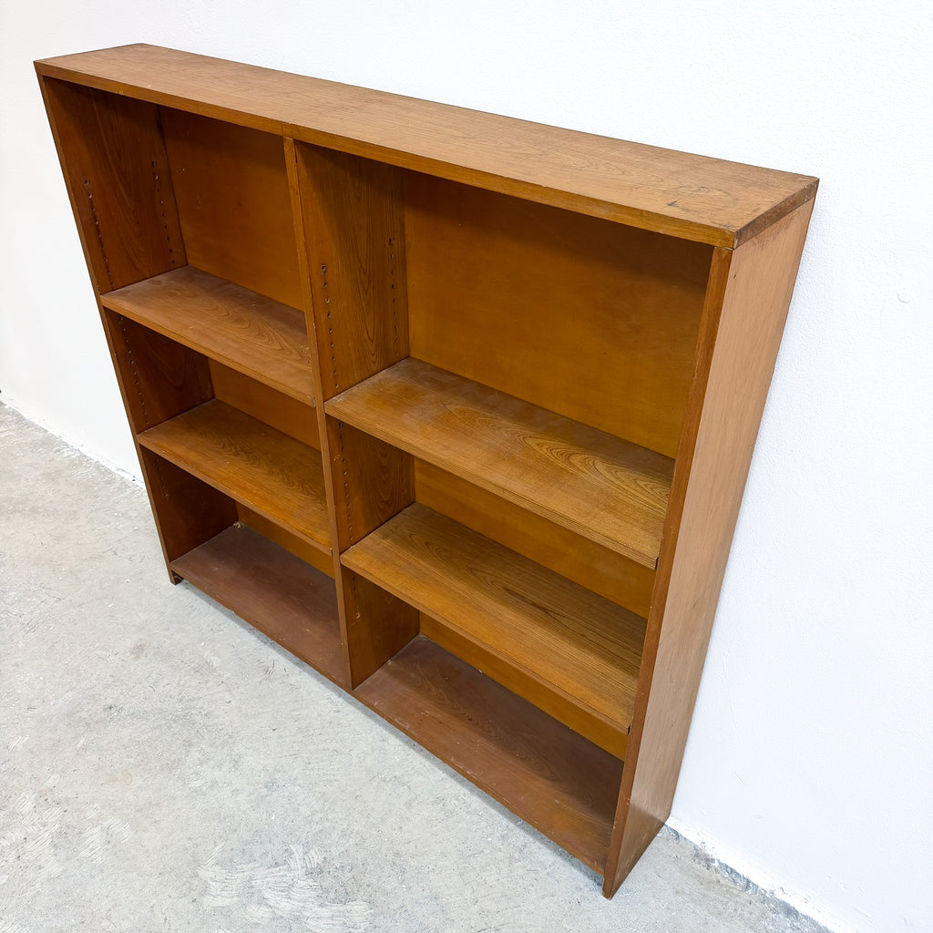 A mid-century modern wooden bookcase with a warm medium-brown finish, featuring six open shelves, photographed against a white wall and concrete floor.
