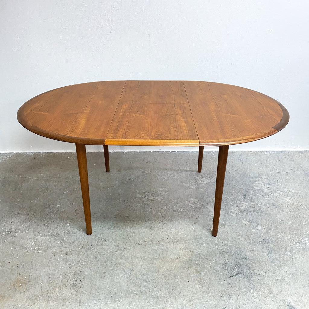 Wooden dining table with a single extension leaf on a concrete floor against a white wall.