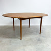 Wooden dining table with a single extension leaf on a concrete floor against a white wall.