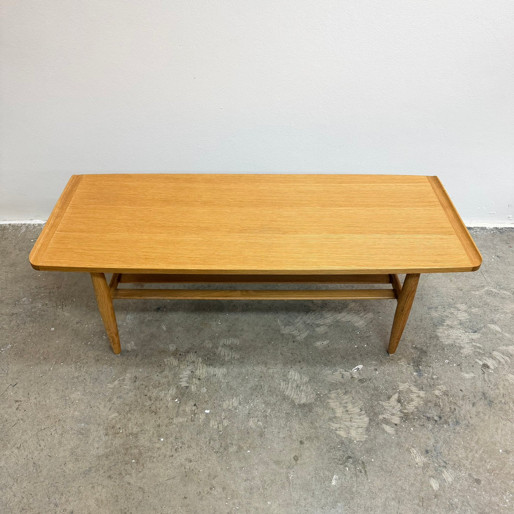 A mid-century modern wooden coffee table with a light oak finish, featuring tapered legs and a lower support rail, set against a plain white wall and concrete floor.
