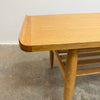 Close-up of a light oak wood table corner showing the rounded edge, wood grain, and part of the slatted base on a concrete floor.