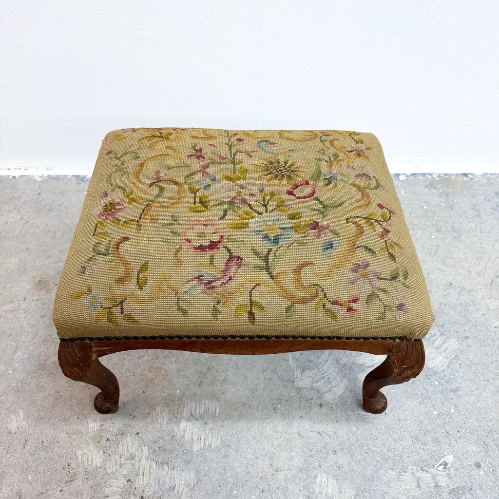 A vintage upholstered wooden stool with a floral needlepoint cushion on a light gray concrete floor against a white wall.