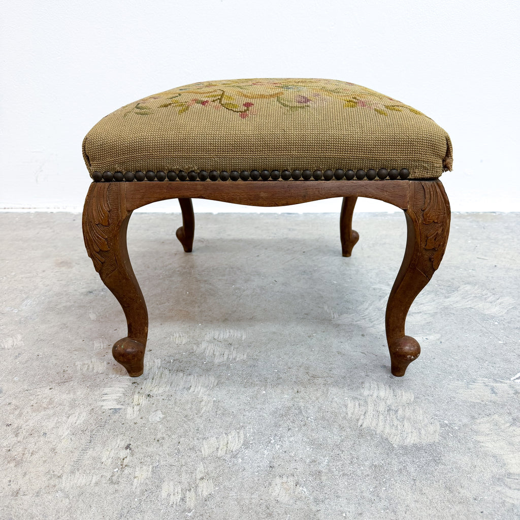 A vintage wooden stool with carved cabriole legs and a tufted needlepoint cushion featuring a floral pattern on a beige background.