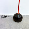 A vintage-style floor lamp with a glossy black spherical base and a bright red vertical stem, standing on a gray concrete floor against a white wall.