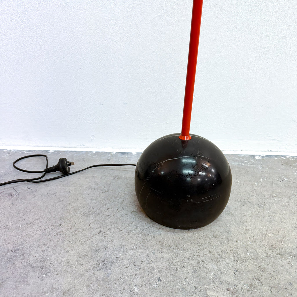 A vintage-style floor lamp with a glossy black spherical base and a bright red vertical stem, standing on a gray concrete floor against a white wall.
