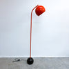 A modern floor lamp with a glossy orange curved arm and dome-shaped shade, mounted on a spherical black base, photographed against a plain white wall and concrete floor.