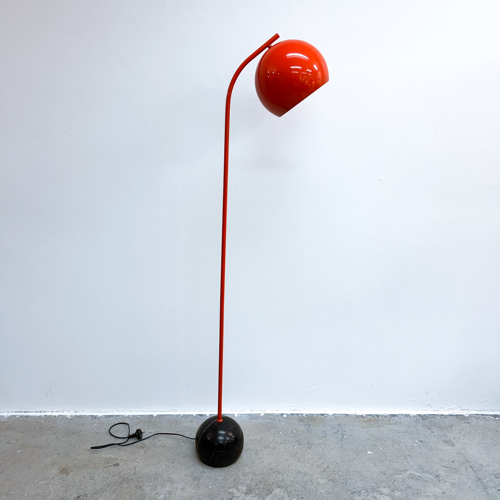 A modern floor lamp with a glossy orange curved arm and dome-shaped shade, mounted on a spherical black base, photographed against a plain white wall and concrete floor.