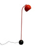 A modern floor lamp with a glossy Orange curved stem, a red spherical shade, and a black spherical base, standing on a concrete floor against a white wall.