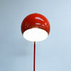 A modern floor lamp with a glossy orange spherical shade and a slim red stem, shown against a pale blue-gray background with the lamp turned on, casting a bright white glow.