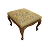 A vintage wooden stool with a floral needlepoint cushion on a concrete floor against a white wall.