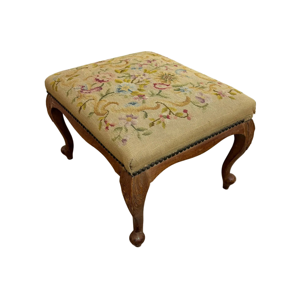 A vintage wooden stool with a floral needlepoint cushion on a concrete floor against a white wall.