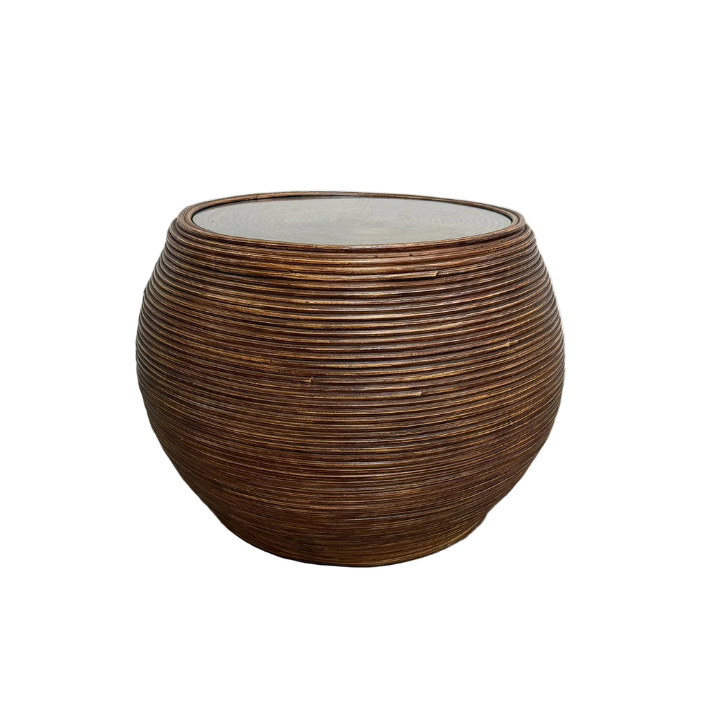 A round, ribbed brown side table with a glass top, set against a plain white wall and gray concrete floor.